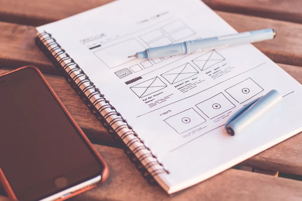 Esboços da estrutura de um website desenhados num caderno, ao lado de um telemóvel e canetas