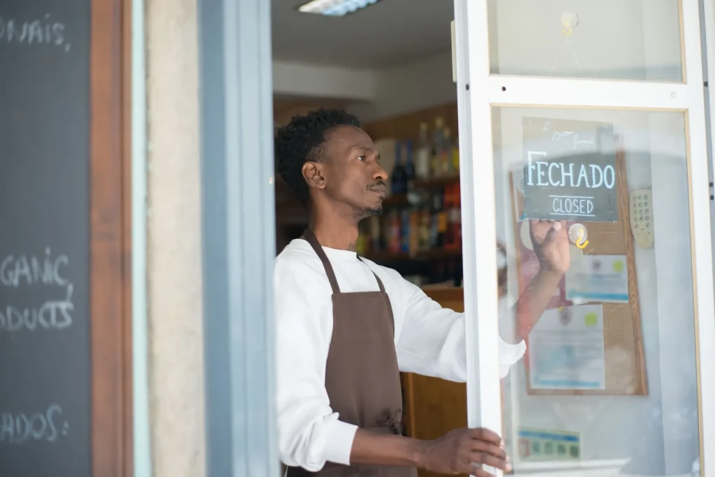 Comerciante à porta de uma pequena loja, ao lado de uma placa com a palavra Fechado